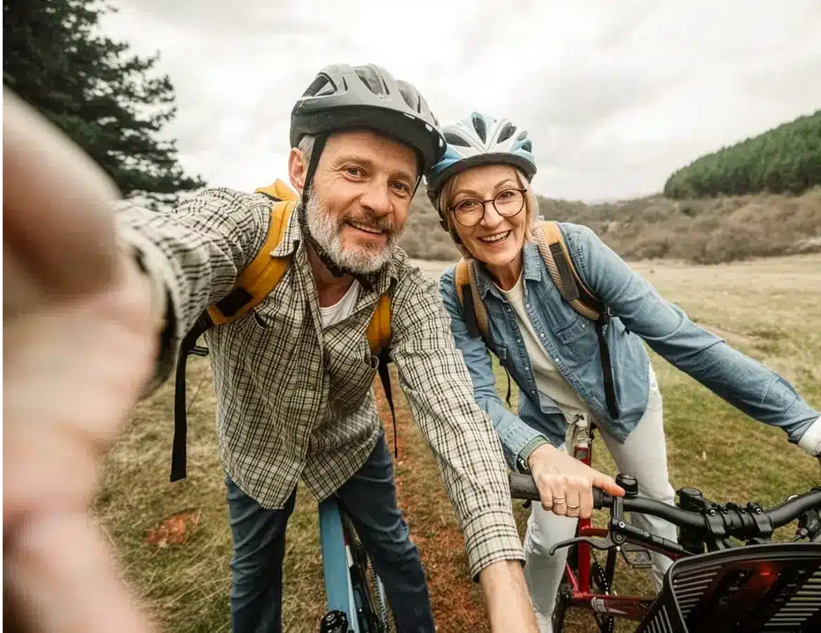 Man and woman wearing bike helmets and backpacks smiling at camera while standing with their bicycles in a grassy field
